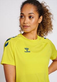 Lyst gul sports skjorte med korte ærmer, som har blå accenter på skuldrene og et logo på brystet. Glat, letvægtsstof.