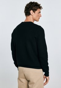 Uomo con capelli ricci di spalle, che indossa un maglione nero a trama e pantaloni beige. Uno sfondo semplice mette in risalto l'outfit.
