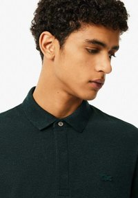 Mörkgrön polo-tröja i texturerad tyg, med knäppbar krage och en liten broderad logotyp på vänster bröst.