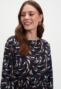 Uśmiechnięta kobieta z długimi brązowymi włosami, nosząca złote kolczyki i granatową sukienkę w białe oraz brązowe abstrakcyjne wzory, patrząca nieco w lewo.