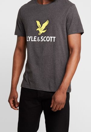 Mężczyzna w ciemnoszarej koszulce Lyle & Scott z żółtym logo orła i czarnych spodniach na jednolitym jasnym tle.