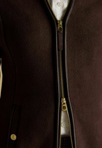 Ciemnobrązowa polarowa kurtka z złotym zamkiem częściowo rozpięta, ukazująca białą koszulę z guzikami pod spodem.