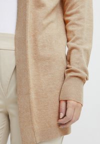 Beige stickad cardigan med ribbad manschetter och nederkant, med en mjuk textur och öppen design framtill. Lämplig för att lagra ovanpå olika outfits.