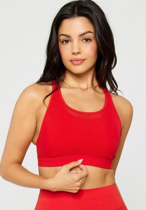 Frau mit langem dunklem Haar, die einen roten Sport-BH und rote hoch taillierte Leggings trägt, posiert vor einem schlichten hellen Hintergrund.