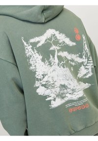 Vihreä huppari pehmeästä kankaasta, jossa on valkoinen maisemakuva ja tekstitulostus takana, punaisilla yksityiskohdilla yläosassa.