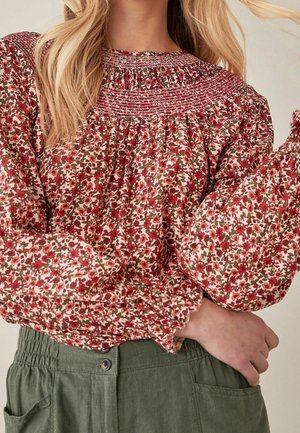 Femme portant un chemisier à manches longues à motifs floraux rouges et roses associé à un pantalon taille haute vert olive avec des boutons.