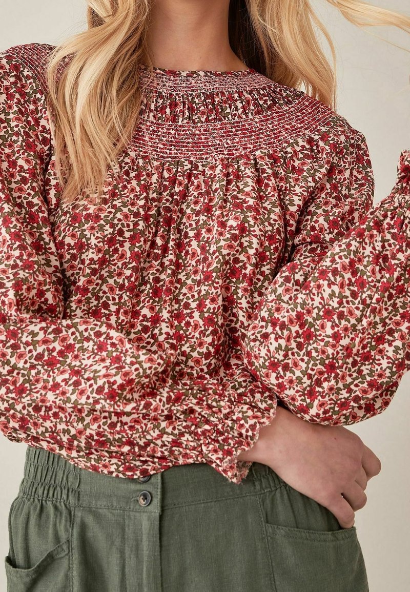 Femme portant un chemisier à manches longues à motifs floraux rouges et roses associé à un pantalon taille haute vert olive avec des boutons.