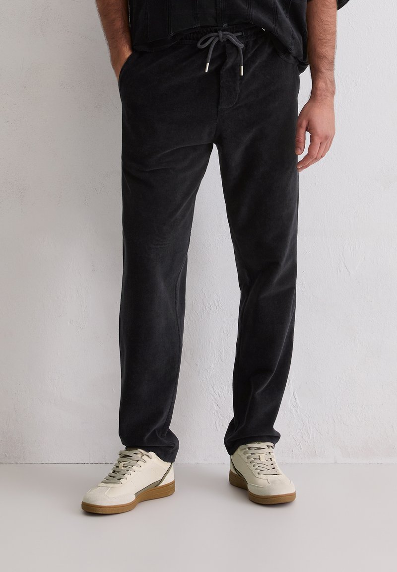 Mann in schwarzen Cordhosen mit Kordelzugbund und beigen Sneakers mit Gummisohlen, der an einer hellen, strukturierten Wand steht.