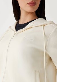 Kremasti zip-up hoodie s kapuljačom, sa reljefnim logom "GUESS". Glatka tekstura i ravni vezice, složen preko tamnoplave majice.