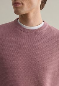 Textured light pink sweater with a round neckline, featuring a subtle patterned weave and layered over a white undershirt.
