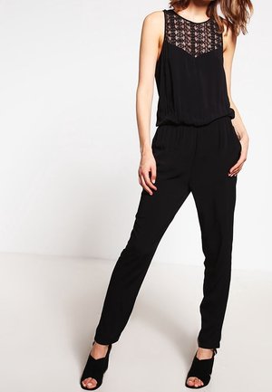 Sort jumpsuit med en blondepanel ved halsudskæringen, uden ærmer, elastisk talje og tætsiddende ben, parret med sorte højhælede sandaler.