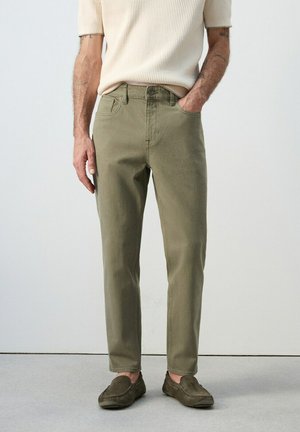 Hombre vistiendo pantalones rectos de color verde oliva, camiseta de punto crema de manga corta y mocasines a juego en verde oliva, de pie con una mano en el bolsillo.