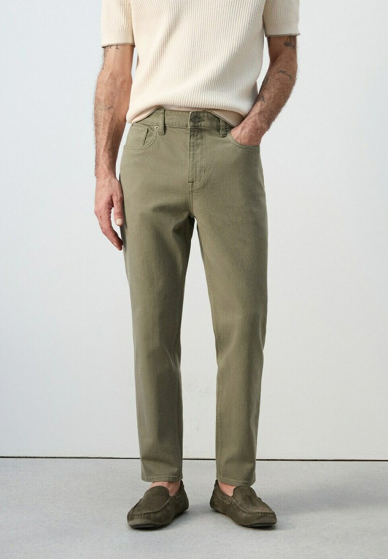 Mann trägt olivgrüne gerade geschnittene Hosen, cremefarbenes kurzärmeliges Strickhemd und passende olivgrüne Loafers, steht mit einer Hand in der Tasche.