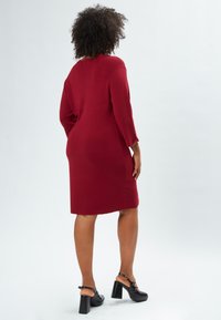 Bordeaux Strickkleid mit 3/4-Ärmeln, gerader Schnitt, bis zur Mitte des Oberschenkels. Getragen mit schwarzen Plateauschuhen, die eine glatte Textur und Passform zeigen.
