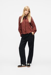 Ξανθιά γυναίκα που στέκεται με τα χέρια στις τσέπες, φορώντας κόκκινο και μαύρο καρό πουκάμισο, μαύρο παντελόνι με φαρδιά μπατζάκια και μαύρα αθλητικά παπούτσια με λευκές σόλες.