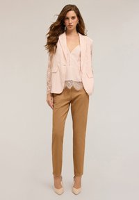 Blazer in pizzo rosa chiaro con maniche testurizzate, indossato sopra un top coordinato. Pantaloni aderenti marroni, completati da tacchi alti beige.