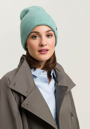 Gebreide beanie in licht teal met een ribbelstructuur; persoon draagt een grijze jas en een lichtblauwe shirt, wat een moderne gelaagde look laat zien.
