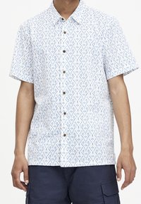 Kortærmet skjorte med knaplukning i hvid bundfarve og blåt geometrisk mønster. Har brune knapper og en standard krave.