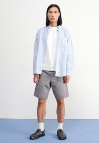 Lyseblå rutete langermet skjorte over en hvit T-skjorte, sammen med grå shorts. Modellen har på seg svarte sko og hvite sokker. Enkelt design.