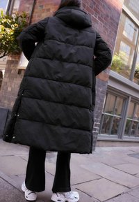 Manteau rembourré noir long avec capuche, présentant un design matelassé et des boutons-pression sur les côtés. Porté avec un pantalon noir évasé et des baskets blanches.