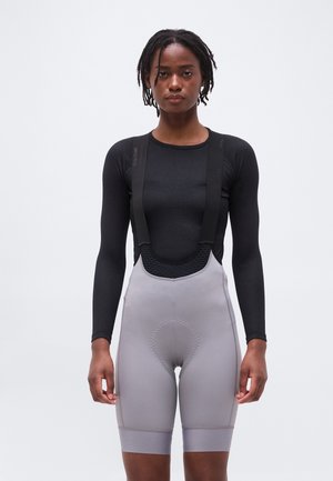 Personne portant un haut noir à manches longues et un cuissard de cyclisme matelassé gris avec bretelles, debout devant un fond clair uni.