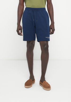 Mężczyzna w granatowych spodenkach z troczkami z małym białym logo na lewej nodze, brązowych butach typu slip-on oraz oliwkowozielonym topie na białym tle.