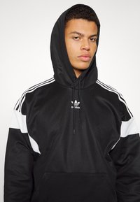 Felpa nera con cappuccio e dettagli bianchi, dotata di tasca a marsupio e tre strisce bianche su ciascun maniche. Logo Adidas sul petto.