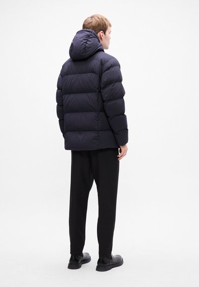 Doudoune marine avec capuche, présentant des sections matelassées et une finition lisse, associée à un pantalon noir et des chaussures.