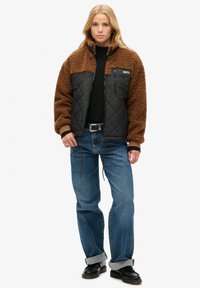 Giacca in pile marrone con pannello trapuntato nero, colletto alto e tasca sul petto con cerniera, abbinata a jeans blu e scarpe nere.