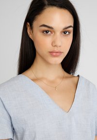 Collana d'oro con la lettera "E", indossata sopra una maglietta azzurra a V realizzata in un tessuto testurizzato. Pelle liscia e capelli scuri visibili.