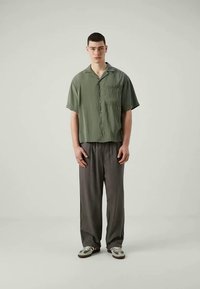 Cămașă cu mânecă scurtă, guler, dintr-un material verde măsliniu, cu o buzunar frontal, asortată cu pantaloni gri lejeri și pantofi casual cu accente verzi.