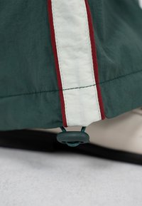 Mørkegrønt stof med en lodret hvid stribe afgrænset af smalle røde linjer og en sort justerbar snorelås i den nederste kant.