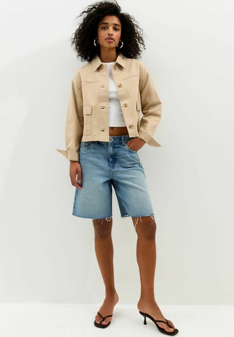 Mujer con cabello rizado que lleva una chaqueta beige abotonada, top corto blanco, shorts largos de mezclilla y sandalias negras de tacón, posando frente a una pared lisa.