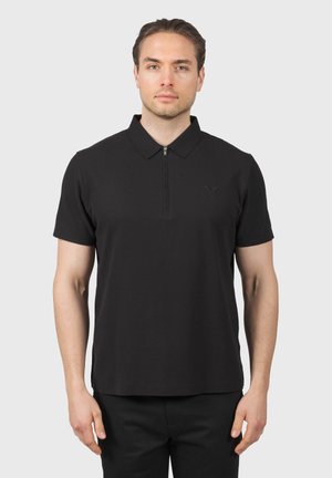 Uomo che indossa una polo nera a maniche corte con una cerniera frontale e colletto, in piedi contro uno sfondo chiaro e uniforme.