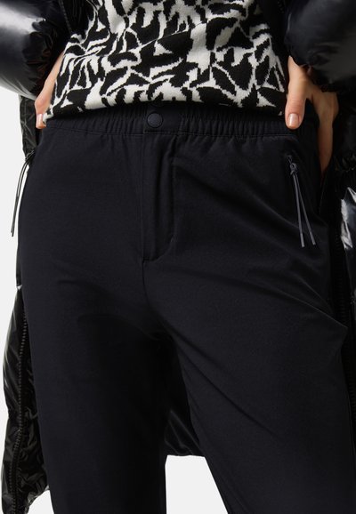 Schwarze, dehnbare Jogginghosen mit elastischem Bund, Seitentaschen und Reißverschlussdetails. Der Stoff hat eine glatte, matte Textur.