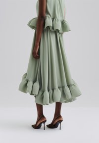 Gaun hijau yang mengalir dengan hem ruffle dan detail peplum, terbuat dari kain ringan. Dipadukan dengan sepatu hak tinggi cokelat.