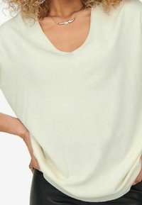 Femme portant un haut crème en V ample, un pantalon noir, une main sur la hanche et un collier en argent avec un pendentif en forme de main.