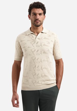 Man draagt een beige korte mouw gebreide polo met abstract patroon, gecombineerd met een donkere broek, staand tegen een effen witte achtergrond.