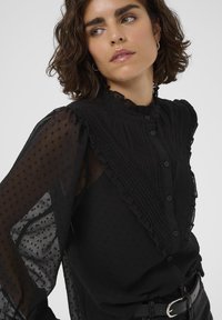 Woman with short curly hair wearing a black sheer blouse with textured dots, ruffles, and buttons, paired with black belt and pants.