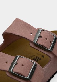 Κοντινό πλάνο ροζ λουριών σανδαλιών από σουέντ με μεταλλικές αγκράφες Birkenstock και φόντο από ταμπά πέλμα.