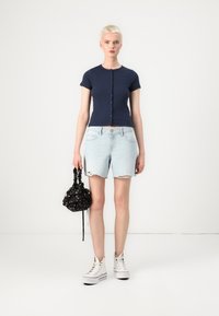 Haut à boutons à manches courtes bleu marine, shorts en denim léger légèrement déchirés, tenant un sac à cordon noir à motifs floraux, associé à des baskets blanches montantes.
