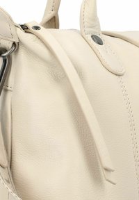 Primo piano di una borsa in pelle beige con cuciture visibili, bottone a scatto nero e una sottile tracolla in pelle che pende vicino all'anello di metallo.