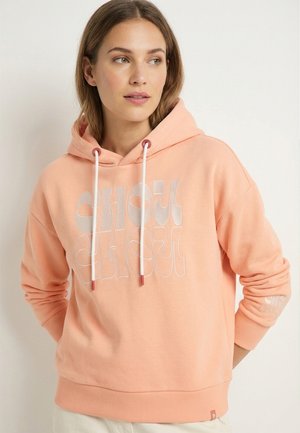 Frau trägt einen pastellpfirsichfarbenen Hoodie mit weißen Kordeln und abstraktem weißen Text, blickt nach links vor einem schlichten, hellen Hintergrund.