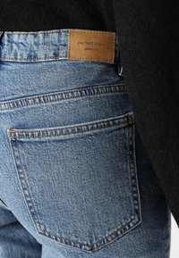 Mėlynos džinsinės kelnės su išblukusiu paviršiumi, turinčios odinį prekės ženklą su užrašu "puikios kelnės" ir dviem užpakaliniais kišenėmis su siuvinėjimu.