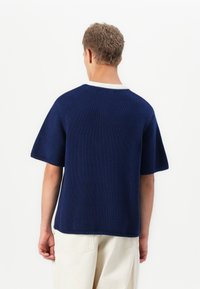 Homme portant un pull en tricot à manches courtes bleu marine avec des bordures blanches, de dos, associé à un pantalon écru sur un fond uni.
