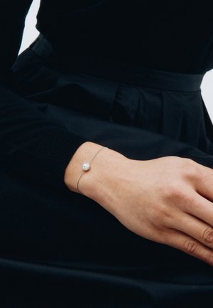 Silbernes Armband mit einem runden Perlenmittelstück, am Handgelenk getragen, vor einem dunklen Stoffhintergrund präsentiert.