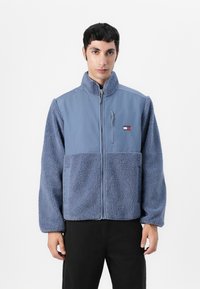 Giacca in pile blu con zip frontale, colletto alto e sezioni in tessuto a contrasto. Include un piccolo logo sul petto e tasche.
