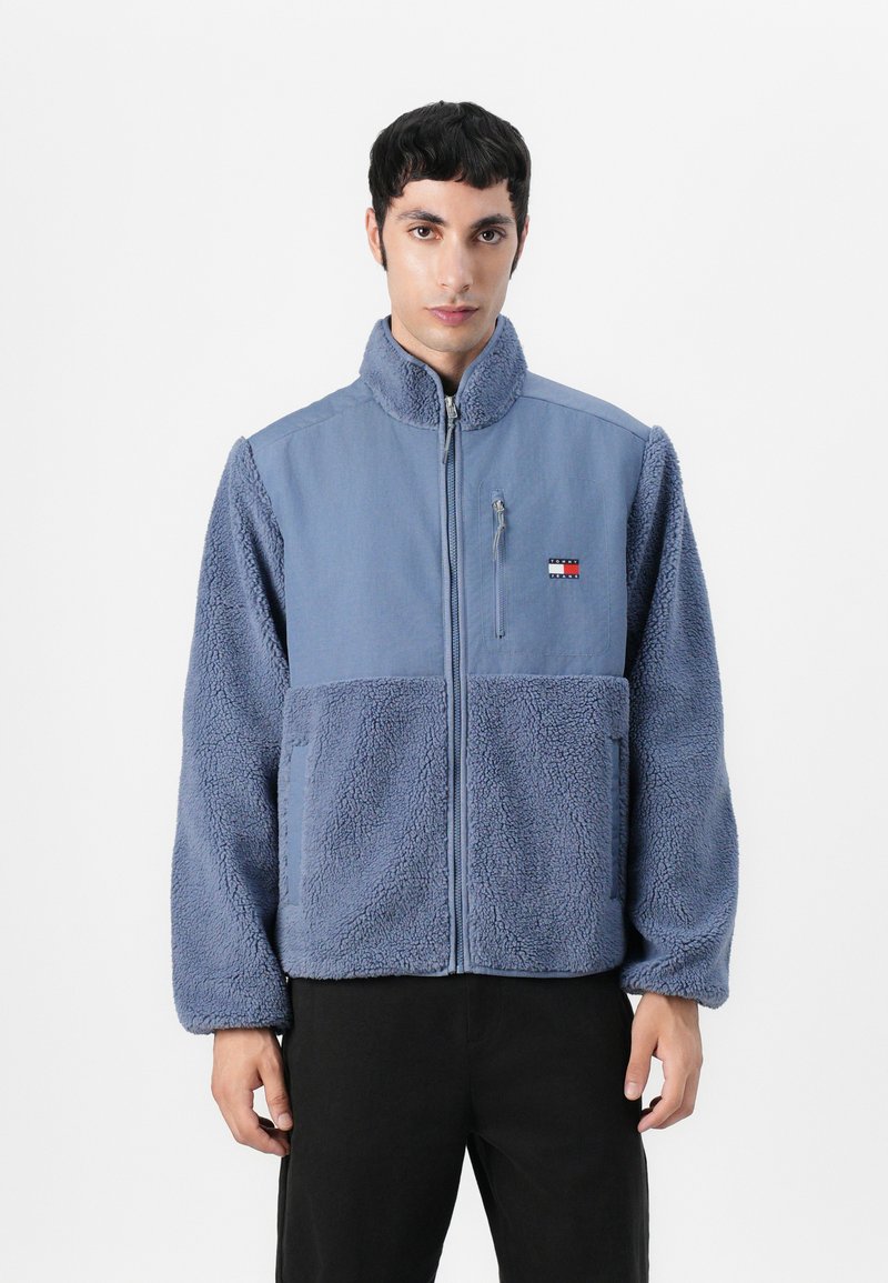 Giacca in pile blu con zip frontale, colletto alto e sezioni in tessuto a contrasto. Include un piccolo logo sul petto e tasche.