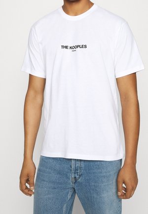 Mężczyzna w białej koszulce z krótkim rękawem z czarnym nadrukiem "THE KOOPLES PARIS" na piersi, w połączeniu z niebieskimi dżinsami.