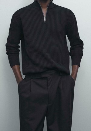 Homme portant un pull côtelé noir avec une demi-fermeture éclair et un pantalon noir, mains dans les poches, sur un fond clair uni.